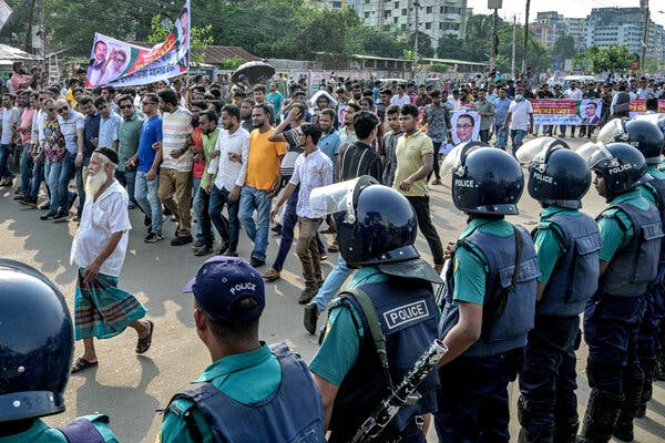 Police officers wearing helmets line the side of a street as a political rally marches by. Some of the marchers are carrying signs with portraits on them. 