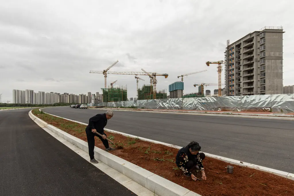 Two people dig in the dirt in median of deserted roadway as construction cranes sit idle in the distance.