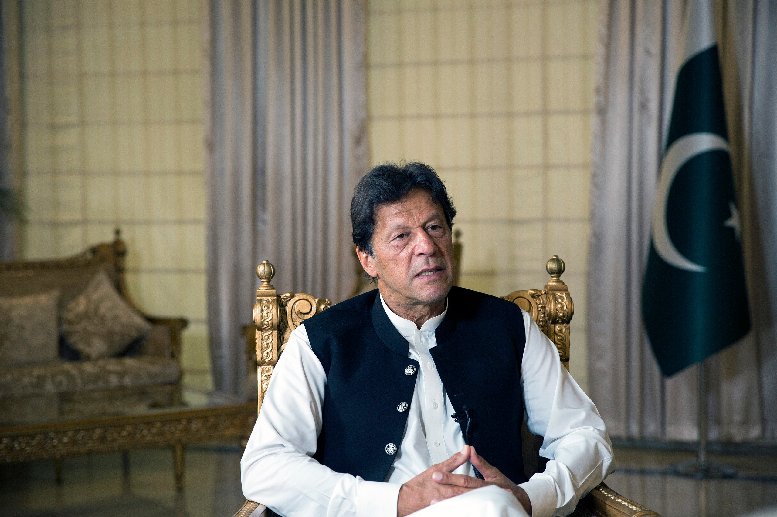 Imran Khan sits in a gold chair with the Pakistani flag behind him his hands crossed in his lap and his mouth open...