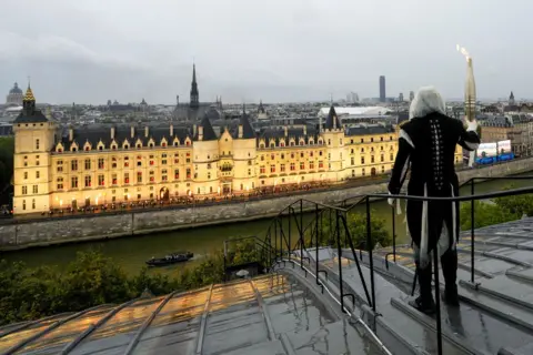  Bernat Armangué/Reuters A hooded and masked torchbearer carries the Olympic flame over a building along the Seine River in Paris, France, during the opening ceremony for the Paris 2024 Olympic Games.