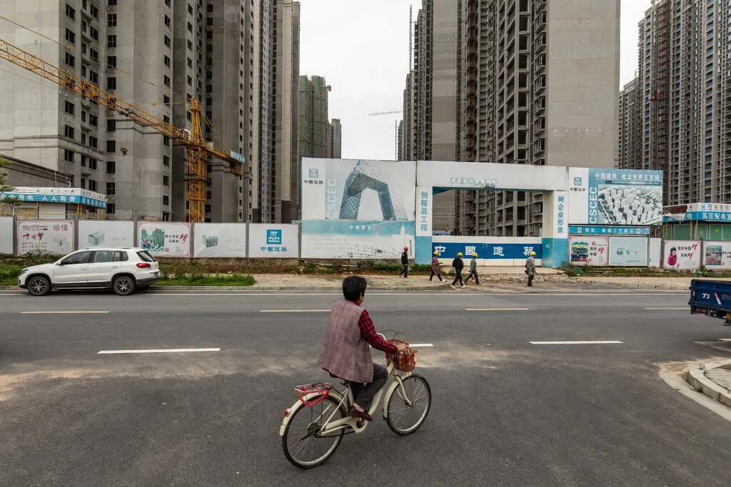 A woman rides a bicycle past a construction site with looming residential towers.