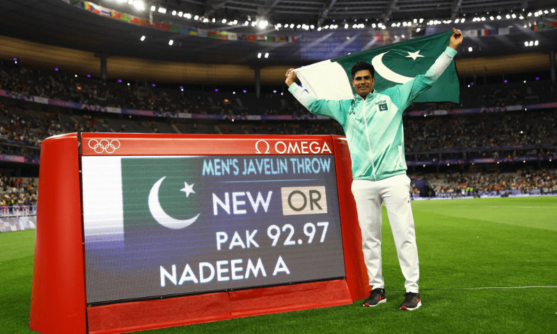 Arshad Nadeem of Pakistan celebrates with his national flag after winning gold as he stands next to a screen displaying his new Olympic record