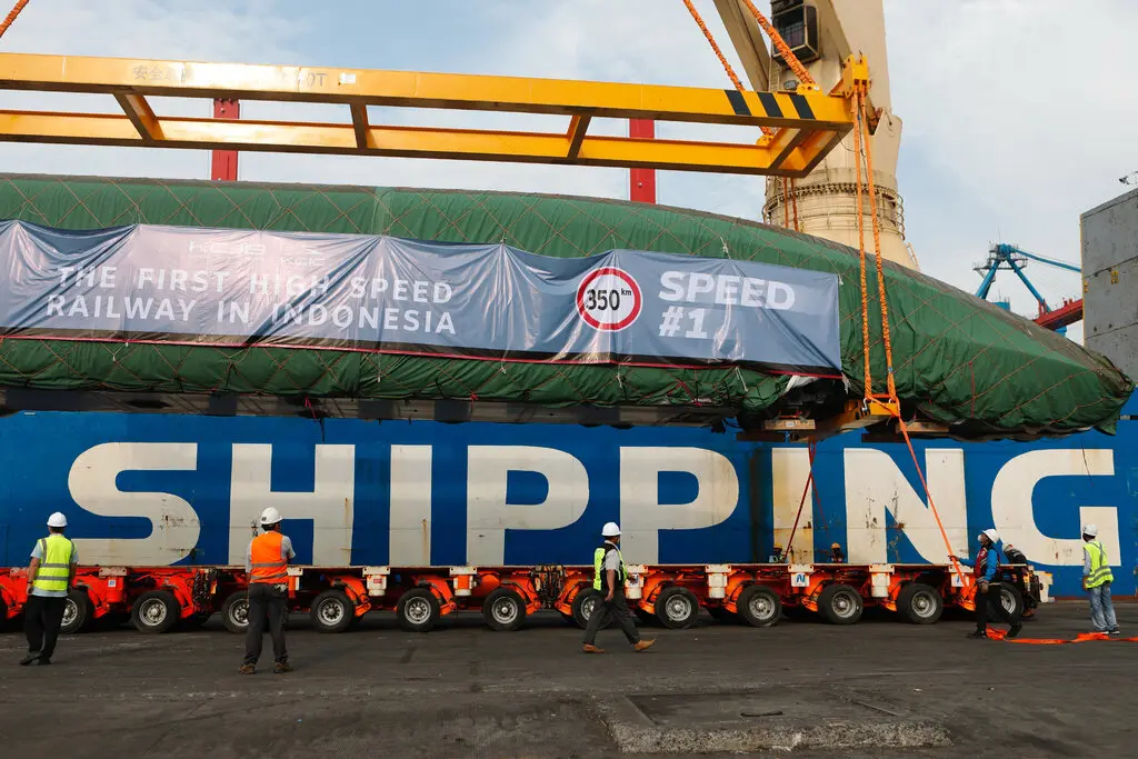 The front of a sleek train is wrapped in green material on a many-wheeled platform that says “Shipping” in capital letter.