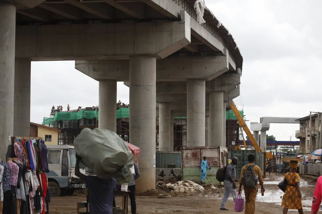 The concrete footings of a highway bridge during construction, with workers on top in the distance and pedestrians beneath it in the foreground.