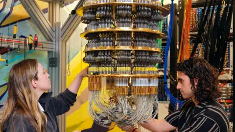 Google Google staff, a woman to the left and a man to the right work on the cryostat which holds the chip and keeps it very cold. The cryostat losley resembles  a chandelier  made of cascading thin metal tubes.